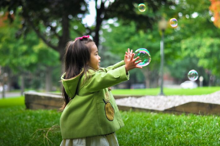 Beautiful photo of young asian child in a park. She wears a green jacket and is playing with bubbles. Her parents have had child custody disputes over her.