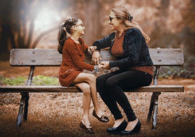 Mother sits in the park in Autumn with her daughter, they are smiling and holding hands. She is talking to her daughter about introducing her daughter to her new partner.